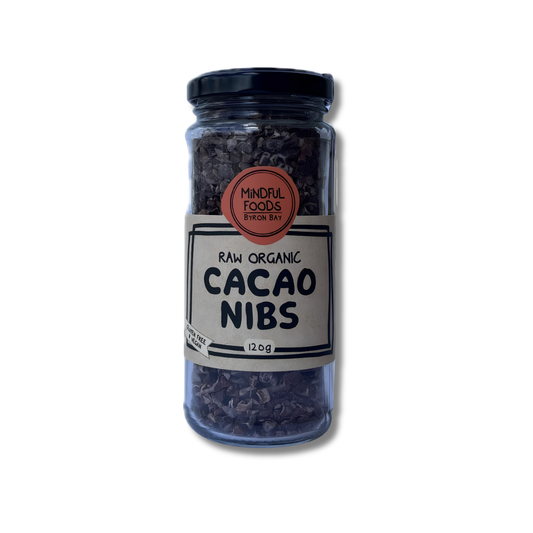 A clear glass jar of Mindful Foods Organic Cacao Nibs, showing the dark brown cacao pieces inside, with a black lid and a white label on a white background.